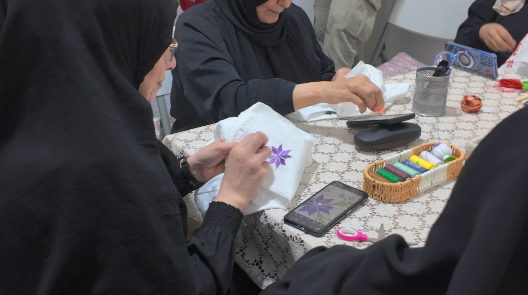 A woman embroiders a pattern on a fabric, tracing the image from a mobile phone.